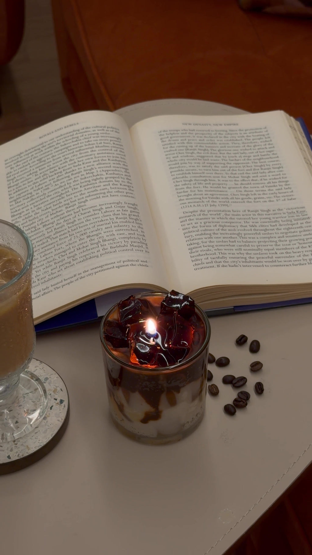 Lit cold coffee candle glowing beside a book and coffee beans